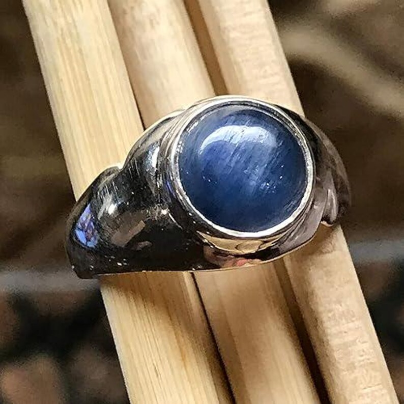 Puede incluir: Un anillo de metal plateado con una piedra preciosa azul de corte cabuj&oacute;n redondo y liso en el centro. La banda tiene una superficie pulida y reflectante que se curva alrededor del dedo, mostrando un tono azul profundo con texturas fibrosas.