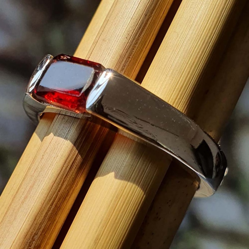 May include: A silver ring with a red gemstone set in a square shape. The ring is on a wooden surface.