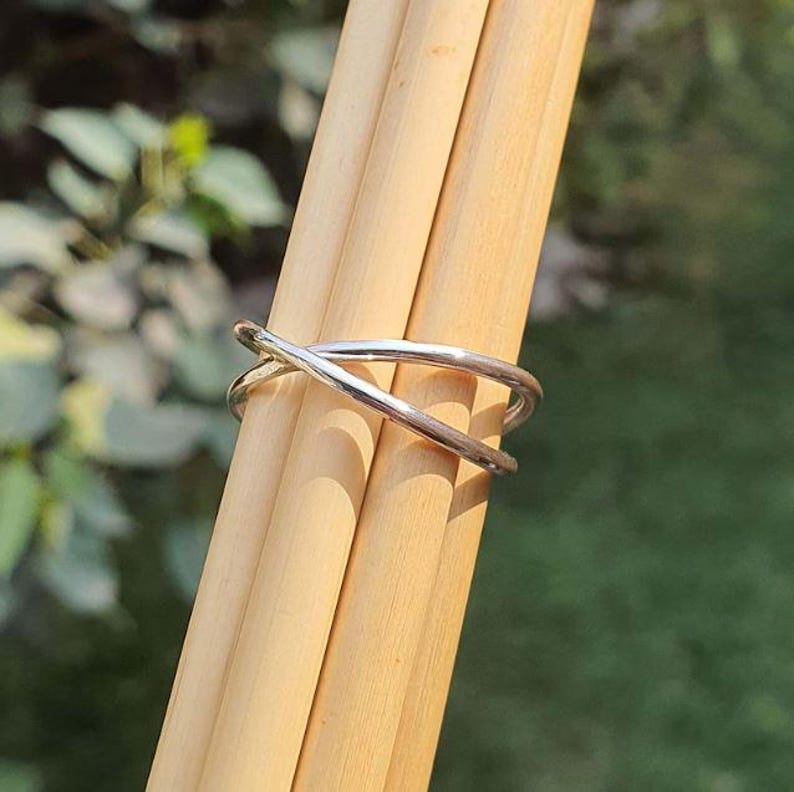 May include: A polished silver-toned ring featuring a crossover design where two thin, smooth bands intersect to form an X shape. The ring is displayed resting on two light-colored wooden dowels, highlighting its reflective metallic surface.