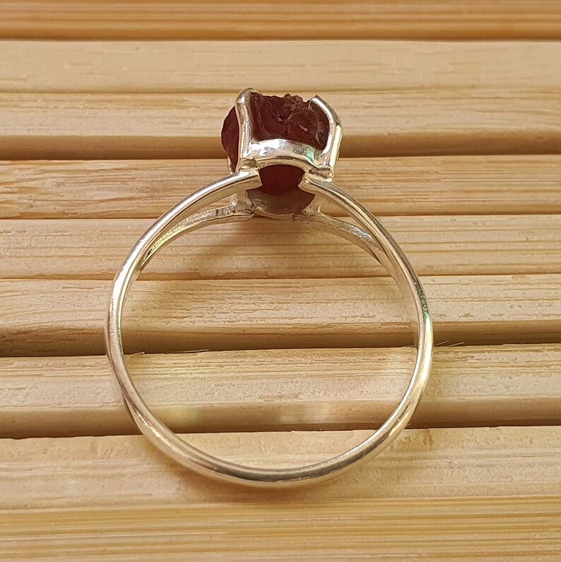 Puede incluir: Un anillo de plata con una piedra preciosa roja engastada en garras. El anillo est&aacute; sobre una superficie de madera.