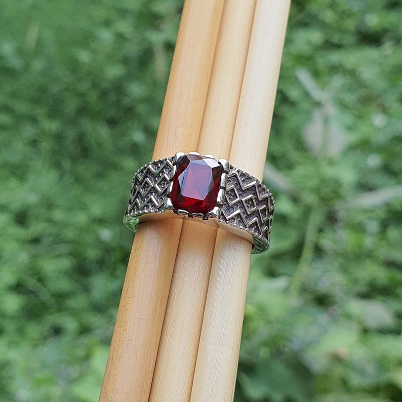 May include: A silver ring with a red gemstone set in a geometric pattern. The ring is on a wooden surface.