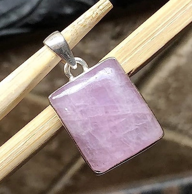 Peut inclure: Un pendentif en argent avec une pierre rectangulaire rose clair. Le pendentif est pos&eacute; sur une surface en bois.