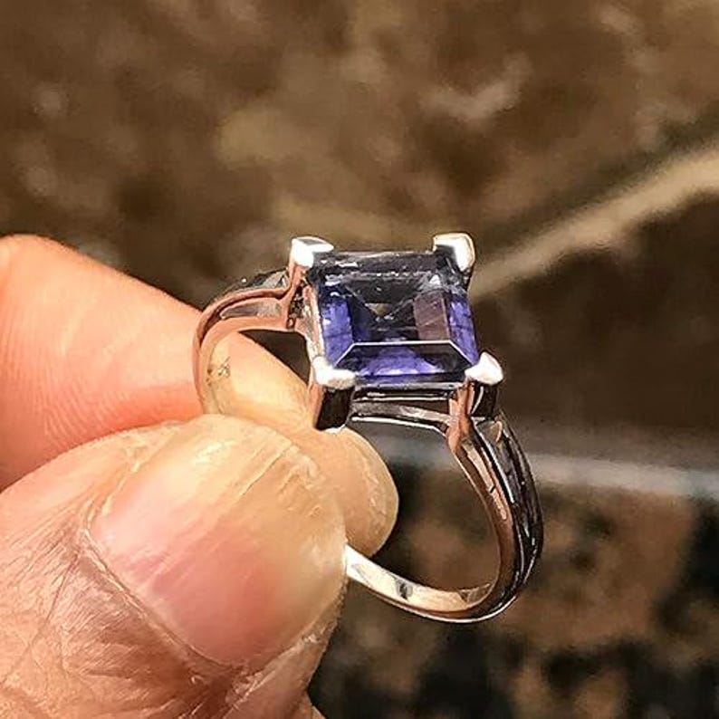 Pode incluir: Um anel de tom prateado com uma pedra preciosa roxa escura de corte quadrado, montada em quatro garras. A banda &eacute; de metal polido e apresenta uma superf&iacute;cie lisa e reflexiva, destacando as facetas da pedra central de forma clara.