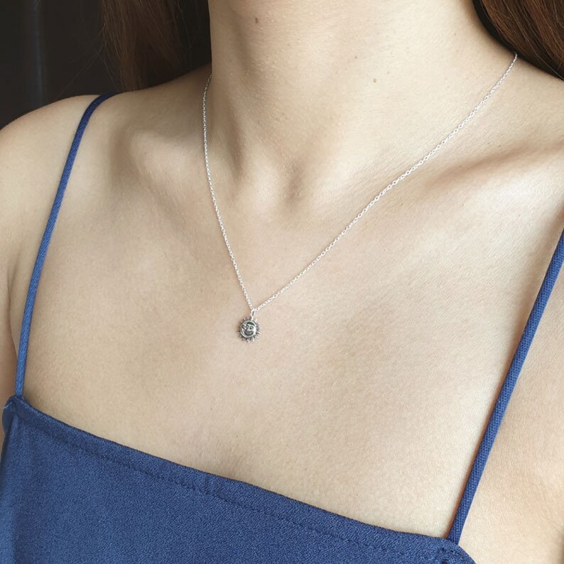 K&ouml;nnte beinhalten: Eine silberfarbene Halskette mit einem kleinen, kreisf&ouml;rmigen Anh&auml;nger, der ein Sonnengesicht zeigt. Der Anh&auml;nger h&auml;ngt an einer zarten, d&uuml;nnen silberfarbenen Kette, die auf dem Schl&uuml;sselbein aufliegt. Das Metall hat ein poliertes Finish.