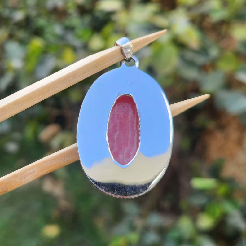 Pode incluir: Pingente de prata com uma pedra oval rosa e branca engastada em uma moldura de prata. O pingente &eacute; segurado por um par de pauzinhos de madeira.
