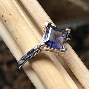 Pode incluir: Um anel de tom prateado com uma &uacute;nica pedra preciosa de cor p&uacute;rpura profunda, de corte quadrado e montada em quatro garras. A banda &eacute; lisa e polida, exibindo um acabamento met&aacute;lico reflexivo que contrasta com a pedra escura facetada.