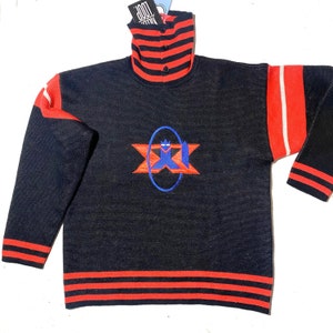 Op de afbeelding: Zwarte trui met rode en witte strepen op de mouwen en de kraag. De trui heeft een groot geborduurd logo op de voorkant met een blauwe cirkel en een rood sterrendesign. Het logo zegt "LOOP NATION".