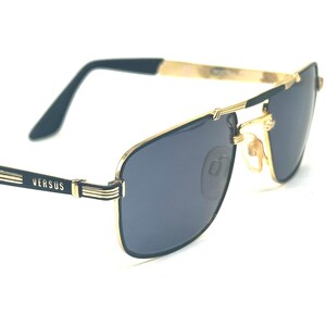 Pu&ograve; includere: Occhiali da sole con montatura dorata e lenti blu scuro. Le montature hanno una forma rettangolare con ponte e aste dorate. Il marchio "VERSUS" &egrave; visibile sull'asta sinistra. Design classico di ispirazione aviator.