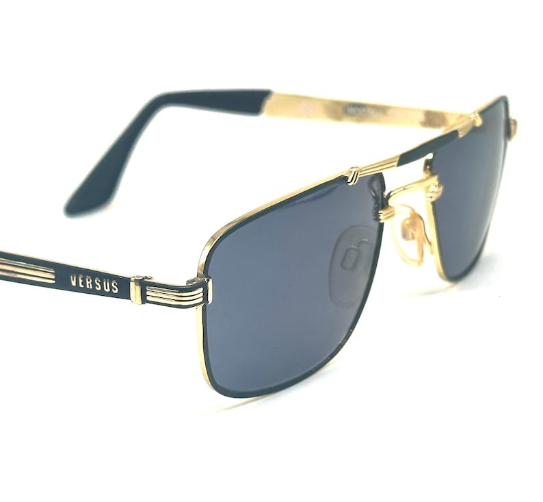 Pu&ograve; includere: Occhiali da sole con montatura dorata e lenti blu scuro. Le montature hanno una forma rettangolare con ponte e aste dorate. Il marchio "VERSUS" &egrave; visibile sull'asta sinistra. Design classico di ispirazione aviator.