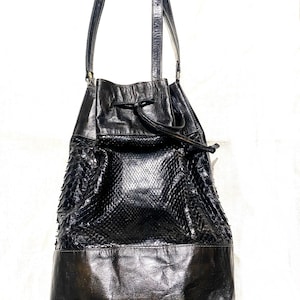 Puede incluir: Un bolso de cubo de cuero negro con un patrón de piel de serpiente y un cierre de cordón. El bolso tiene una correa larga y un fondo hecho de un tipo diferente de cuero.