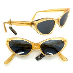 Puede incluir: Gafas de sol vintage de ojo de gato con montura ámbar claro y lentes oscuras. Las monturas tienen un patrón decorativo. Las gafas de sol tienen una etiqueta que dice "MADE IN ITALY".
