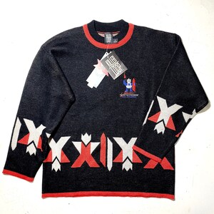 Op de afbeelding: Zwarte gebreide trui met rode en witte geometrische patronen. De trui heeft een rode ronde hals en een klein logo-patch aan de voorkant met de tekst "Killer Loop".