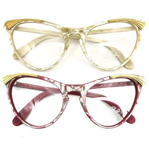 Op de afbeelding: Twee paar cat-eye brillen. Eén paar heeft een crèmekleurig montuur met gouden accenten en het andere een bordeauxrood montuur met gouden accenten. Beide paren hebben heldere glazen en de merknaam "Retro Metalvista" is zichtbaar.