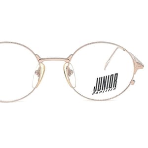 May include: Rose gold round eyeglasses with a Junior Gaultier logo on the right lens.