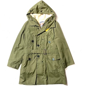 Op de afbeelding: Olijfgroene parka met capuchon en imitatiebontvoering. De jas heeft een ceintuur in de taille, meerdere zakken en knoopsluitingen. Een geel label hangt aan de jas. De capuchon heeft drukknoopsluitingen. De jas is in vintage stijl.