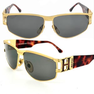 May include: Gold-framed sunglasses with dark gray lenses. The sunglasses have a rectangular shape with a gold bridge and temples. The temples feature a brown tortoiseshell pattern and the letter "H" with the brand name "HILTON".