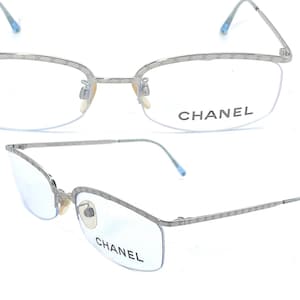 May include: Silver-toned Chanel eyeglasses with a semi-rimless design. The frames feature the Chanel logo along the top edge and the brand name is printed on the lens. The temples and tips are a light blue color.
