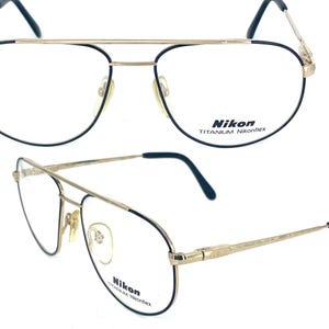 Puede incluir: Gafas Nikon Titanium Nikonflex doradas con doble puente y detalles en negro. Las lentes son de forma ovalada con una montura negra fina. Las patillas son finas y doradas. El logotipo de Nikon es visible en las lentes y las patillas.