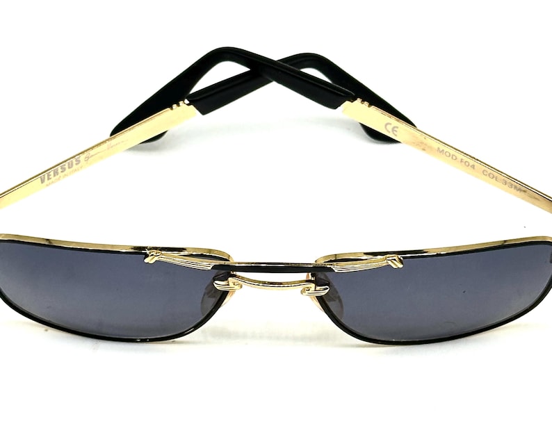 Pu&ograve; includere: Occhiali da sole con montatura dorata e lenti grigio scuro. Gli occhiali da sole hanno un design elegante e moderno con aste nere e il marchio "VERSUS" visibile sulla montatura. Le lenti sono di forma curva e semi-senza montatura.