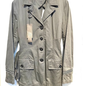 Puede incluir: Una chaqueta gris de manga larga con cinturón, dos bolsillos y botones negros. La chaqueta tiene rayas blancas en los puños. La etiqueta de la chaqueta dice "J.P. GAIA" y "PROJECTS".