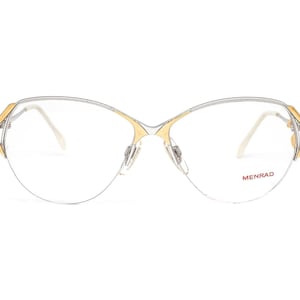 May include: Silver and gold-toned eyeglasses with a cat-eye shape and clear lenses. The brand name "MENRAD" is printed in red on the right lens.