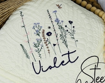 May include: A white quilted blanket with a floral embroidery design and the name "Violet" embroidered in navy blue. The blanket has a fringe trim and is decorated with wooden beads.