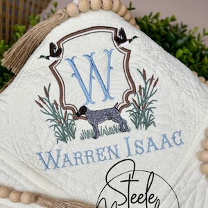 May include: A white quilted blanket with a brown and blue embroidered design featuring a dog, birds, and the letter W. The blanket has a brown tassel and is surrounded by wooden beads. The text "Warren Isaag" and "Steele Magnolia Custom Embroidery and Gifts" is embroidered on the blanket.