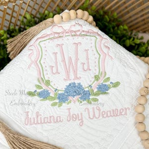 May include: White quilted baby blanket with pink and green embroidered floral design and the name "Juliana Joy Weaver". Features a monogram and tassel accents. A decorative item for a nursery.