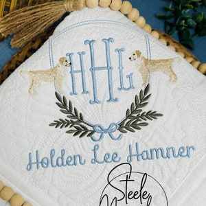 May include: White quilted baby blanket with a blue and green embroidered monogram, two golden retriever dogs, and the name "Holden Lee Hammer". The blanket has a fringe trim and is decorated with wooden beads. The bottom right corner of the blanket has a logo for "Steele Magnolia Custom Embroidery and Gifts".