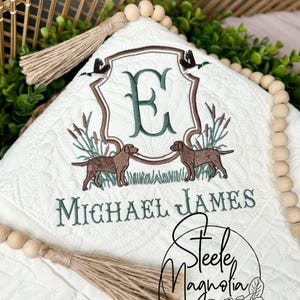 May include: Cream-colored, quilted blanket with embroidered details. Features a central crest with the letter "E", two dogs, and the name "Michael James". Includes a decorative tassel and wooden beads. The blanket is adorned with images of ducks and cattails.