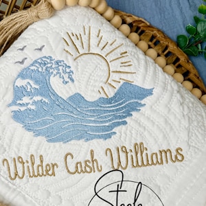 May include: White quilted baby blanket with a blue wave, sun, and birds embroidered on it. The blanket also has the name "Wilder Cash Williams" embroidered in gold thread. The blanket is decorated with a brown tassel and wooden beads.