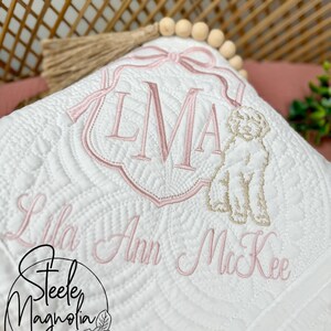 K&ouml;nnte beinhalten: Wei&szlig;e gesteppte Babydecke mit rosa Stickerei. Die Decke zeigt ein Monogramm "LMA" mit Schleife, eine Hundefigur und den Namen "Lila Ann McKee".