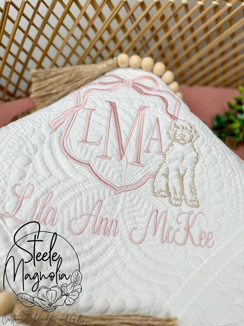 K&ouml;nnte beinhalten: Wei&szlig;e gesteppte Babydecke mit rosa Stickerei. Die Decke zeigt ein Monogramm "LMA" mit Schleife, eine Hundefigur und den Namen "Lila Ann McKee".