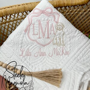 K&ouml;nnte beinhalten: Eine wei&szlig;e gesteppte Babydecke mit einem rosa gestickten Monogramm "LMA" und dem Namen "Lila Ann McKee" in Kursivschrift. Ein kleiner gestickter Hund befindet sich neben dem Monogramm. Die Decke liegt in einem Weidenkorb mit einem braunen Quasten und Holzperlen.