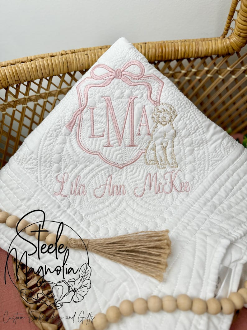 K&ouml;nnte beinhalten: Eine wei&szlig;e gesteppte Babydecke mit einem rosa gestickten Monogramm "LMA" und dem Namen "Lila Ann McKee" in Kursivschrift. Ein kleiner gestickter Hund befindet sich neben dem Monogramm. Die Decke liegt in einem Weidenkorb mit einem braunen Quasten und Holzperlen.