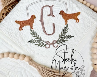 Puede incluir: Una manta de tiro acolchada blanca con una inicial bordada "C" en marrón, rodeada de una corona de hojas verdes y dos perros golden retriever bordados. La manta está decorada con una borla marrón y una guirnalda de cuentas de madera.