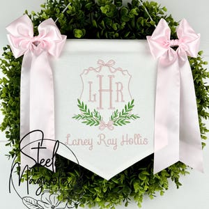 May include: A white banner with pink embroidery, featuring the initials "LHR" within a shield design, surrounded by greenery and bows. The banner is adorned with pink satin bows and the name "Laney Ray Hollis". The banner is set against a green wreath.