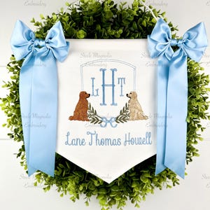 May include: A white banner with blue embroidery, featuring the initials "LHT" and two dog illustrations, is suspended within a green wreath. Light blue bows adorn the sides. The text "Lane Thomas Howell" is also embroidered.