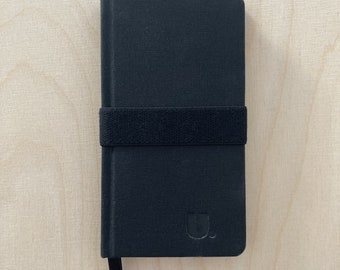 Puede incluir: Un cuaderno negro con cierre de banda elástica negra y un pequeño logotipo negro en la parte delantera.