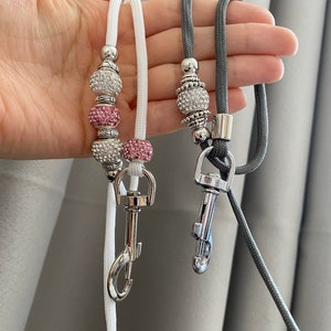 K&ouml;nnte beinhalten: Zwei Perlenb&auml;nder mit silbernen Verschl&uuml;ssen. Ein Band ist wei&szlig; mit rosa und klaren Perlen. Das andere Band ist grau mit klaren Perlen.