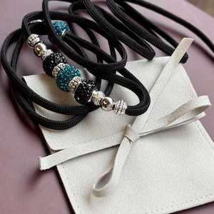 K&ouml;nnte beinhalten: Schwarze Paracord-Hundeleine mit t&uuml;rkisfarbenen und schwarzen Kristallperlen und silbernen Akzenten. Die Leine ist aufgerollt und wird mit einem wei&szlig;en Wildlederbeutel geliefert.