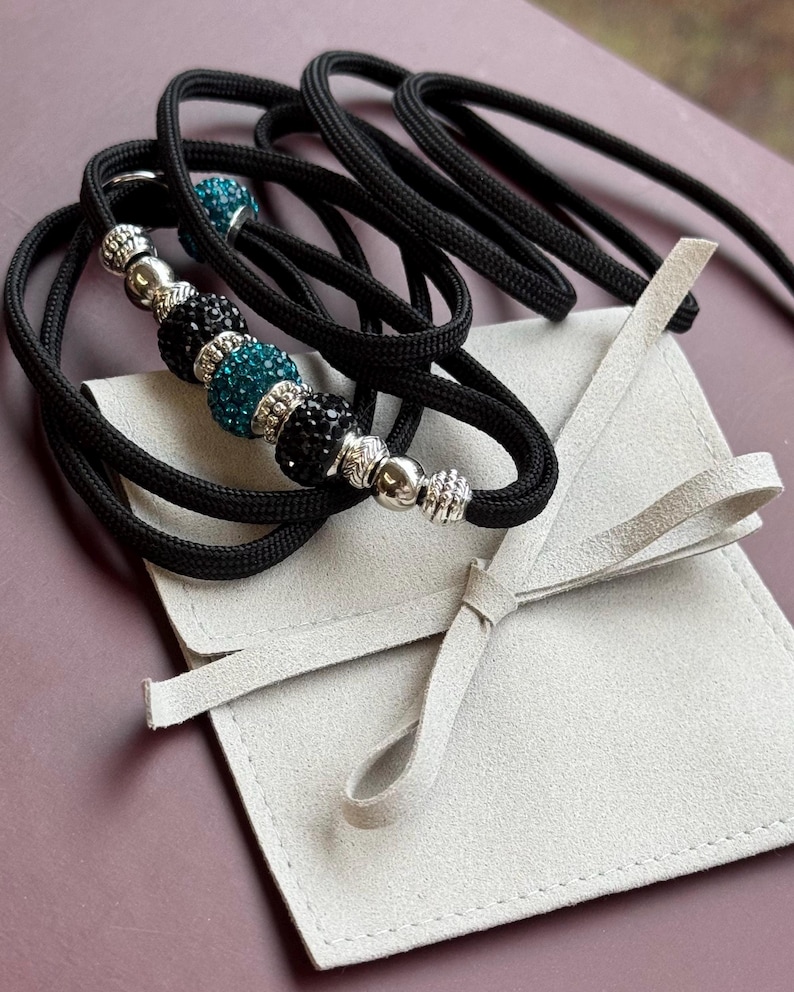 K&ouml;nnte beinhalten: Schwarze Paracord-Hundeleine mit t&uuml;rkisfarbenen und schwarzen Kristallperlen und silbernen Akzenten. Die Leine ist aufgerollt und wird mit einem wei&szlig;en Wildlederbeutel geliefert.