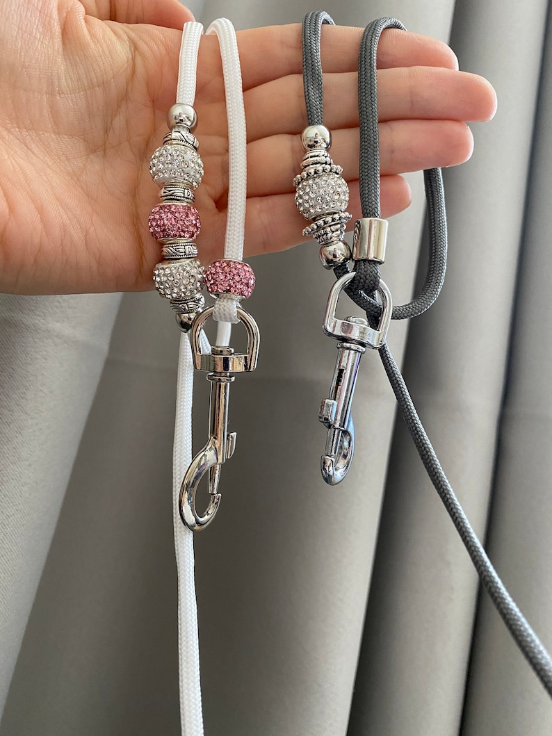 K&ouml;nnte beinhalten: Zwei Perlenb&auml;nder mit silbernen Verschl&uuml;ssen. Ein Band ist wei&szlig; mit rosa und klaren Perlen. Das andere Band ist grau mit klaren Perlen.