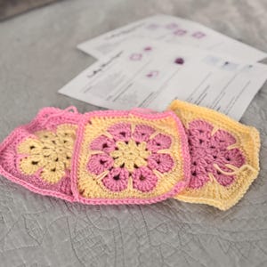 May include: Three handmade crochet squares in pink and yellow, arranged on a grey surface. Each square features a floral design in the centre. Crochet pattern instructions are visible in the background.