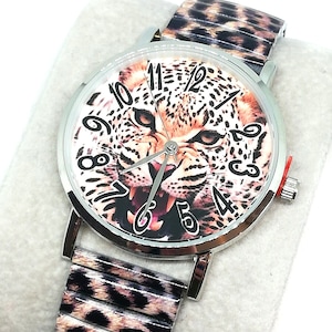 Könnte beinhalten: Eine silberfarbene Armbanduhr mit Leopardenmuster-Armband und Zifferblatt. Das Zifferblatt zeigt eine Nahaufnahme des Gesichts eines Leoparden mit den Zahlen 1 bis 12. Das Armband hat ein passendes Leopardenmuster. Die Uhr liegt auf einer weißen Oberfläche.