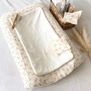 Op de afbeelding: Een witte verschoningsmat met een bloemenpatroon, een witte handdoek, een houten borstel en een rieten mand gevuld met babybenodigdheden.