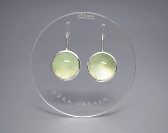 May include: A pair of round, light green cabochon earrings set in silver-toned metal with hook backings. The earrings are displayed on a clear circular acrylic stand featuring the etched text "VERBA" at the top and "AGRA VASARA" at the bottom.