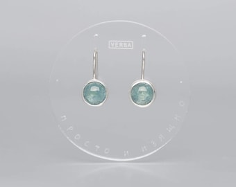 May include: A pair of silver drop earrings with round, light blue gemstones. The earrings are displayed on a clear, circular stand with the word "VERBA" printed at the top.
