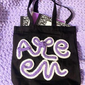 以下が含まれることがあります： 紫と白のかぎ針編みで「ARE EM」の文字が入った黒いトートバッグ。バッグには黒い持ち手があり、ノートが入っています。背景は、薄紫色のテクスチャード加工された毛布です。