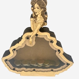 May include: Wooden princess-shaped coin bank. The bank features a laser-cut design of a woman in a ball gown. The bank is made of layered wood, with a clear front panel to view the coins inside. The princess figure is at the top.